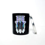 The Zodiac: Gemini Candle is a black jar labeled "Gemini," featuring an illustrated Gemini figure and symbol. It sits on a white background with its partially open silver lid resting beside it.