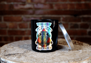 The Zodiac: Aquarius Candle, made from natural soy, sits on a wooden surface with its vibrant zodiac label visible and the silver lid placed nearby.
