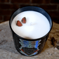 The Zodiac: Leo Candle, a black jar labeled "Leo," features all-natural soy white wax and two small red stones on top, displayed on a round rustic surface.