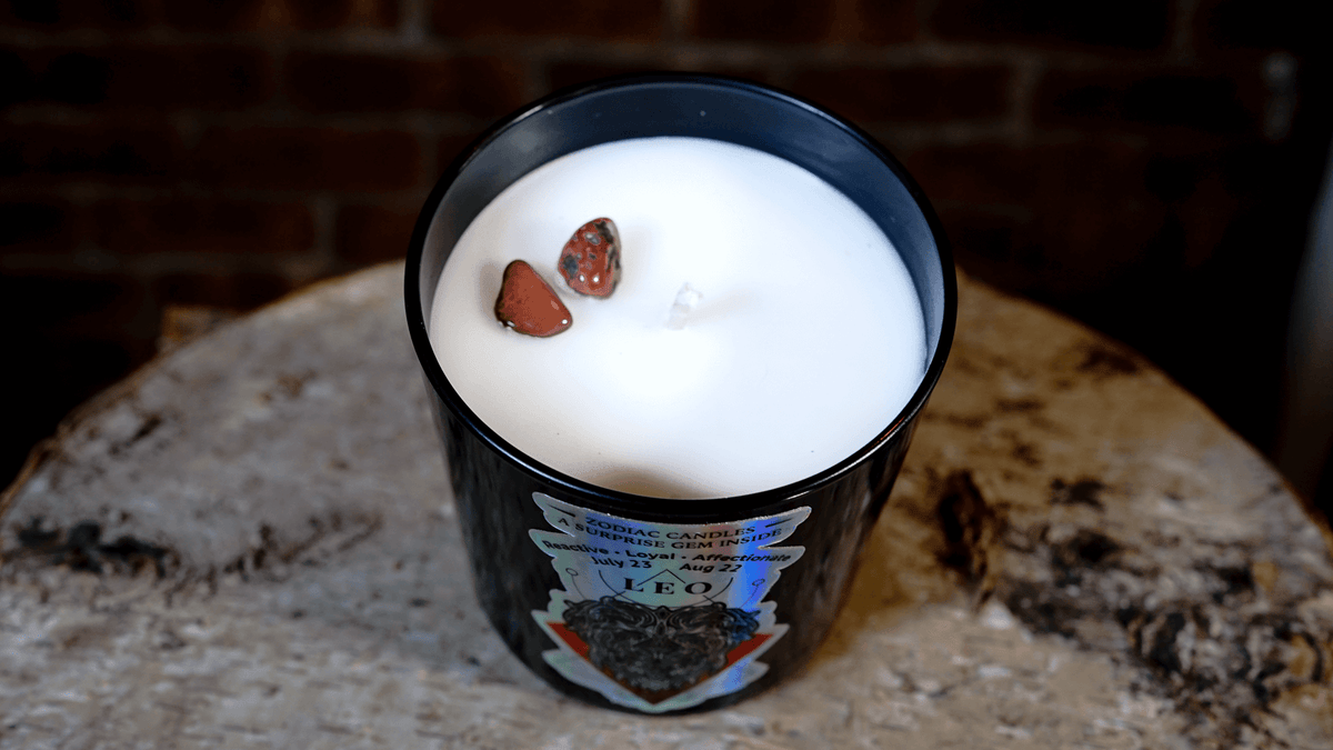 The Zodiac: Leo Candle, a black jar labeled "Leo," features all-natural soy white wax and two small red stones on top, displayed on a round rustic surface.