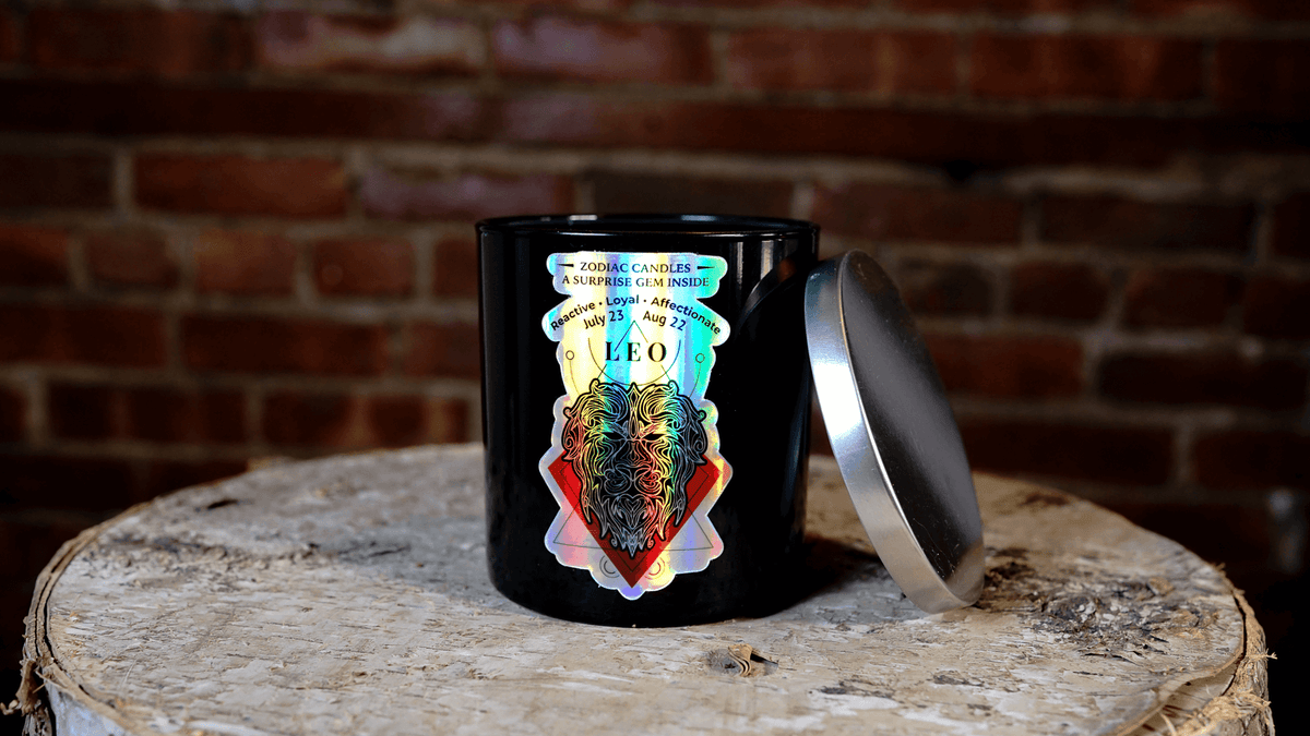 A black, all-natural soy candle jar with a metallic lid labeled “The Zodiac: Leo Candle” sits on a rustic wooden surface in front of a brick wall.