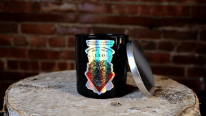 A black, all-natural soy candle jar with a metallic lid labeled “The Zodiac: Leo Candle” sits on a rustic wooden surface in front of a brick wall.
