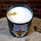 The Zodiac: Sagittarius Candle, a black soy zodiac candle, rests on a rough wooden surface with two yellow stones on top, set against a rustic brick wall.