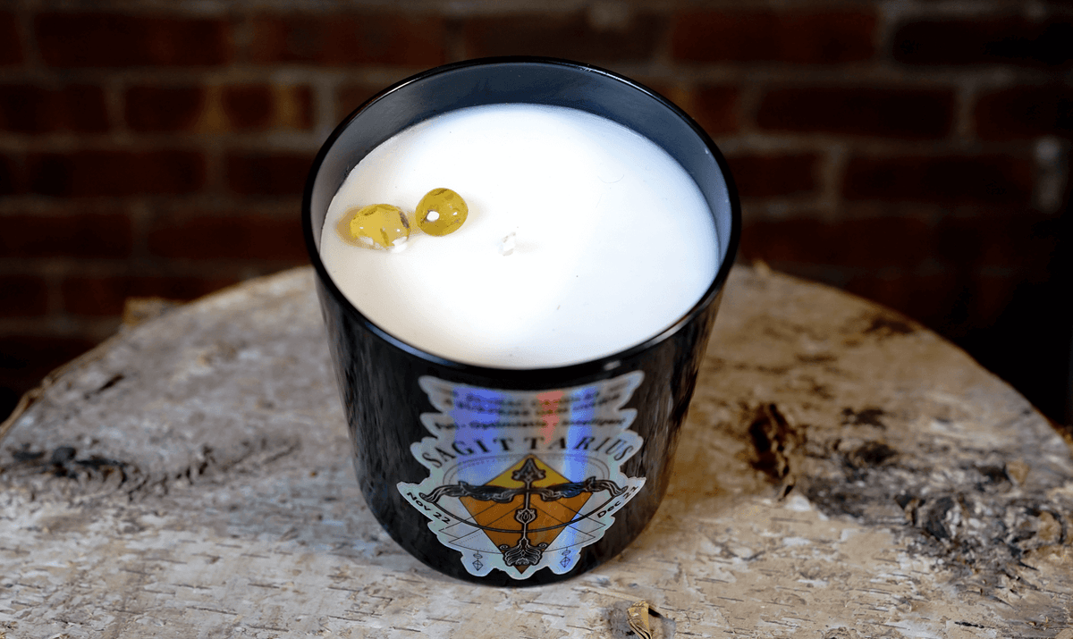 The Zodiac: Sagittarius Candle, a black soy zodiac candle, rests on a rough wooden surface with two yellow stones on top, set against a rustic brick wall.