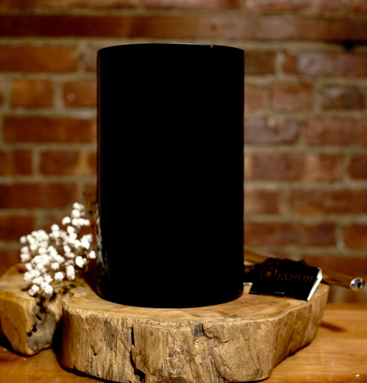 The “Thanks for Puttin’ Up with My Shit” large black soy candle, made from a recycled wine bottle, rests on a wooden slab with dried white flowers and a metal wick trimmer against a brick wall backdrop.