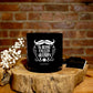 The "Happiness is Being Called Grandpa" Natural Soy Candle sits on a wooden surface, making an ideal gift for grandpa. White flowers and a matchbox lie nearby, all against a brick wall backdrop.
