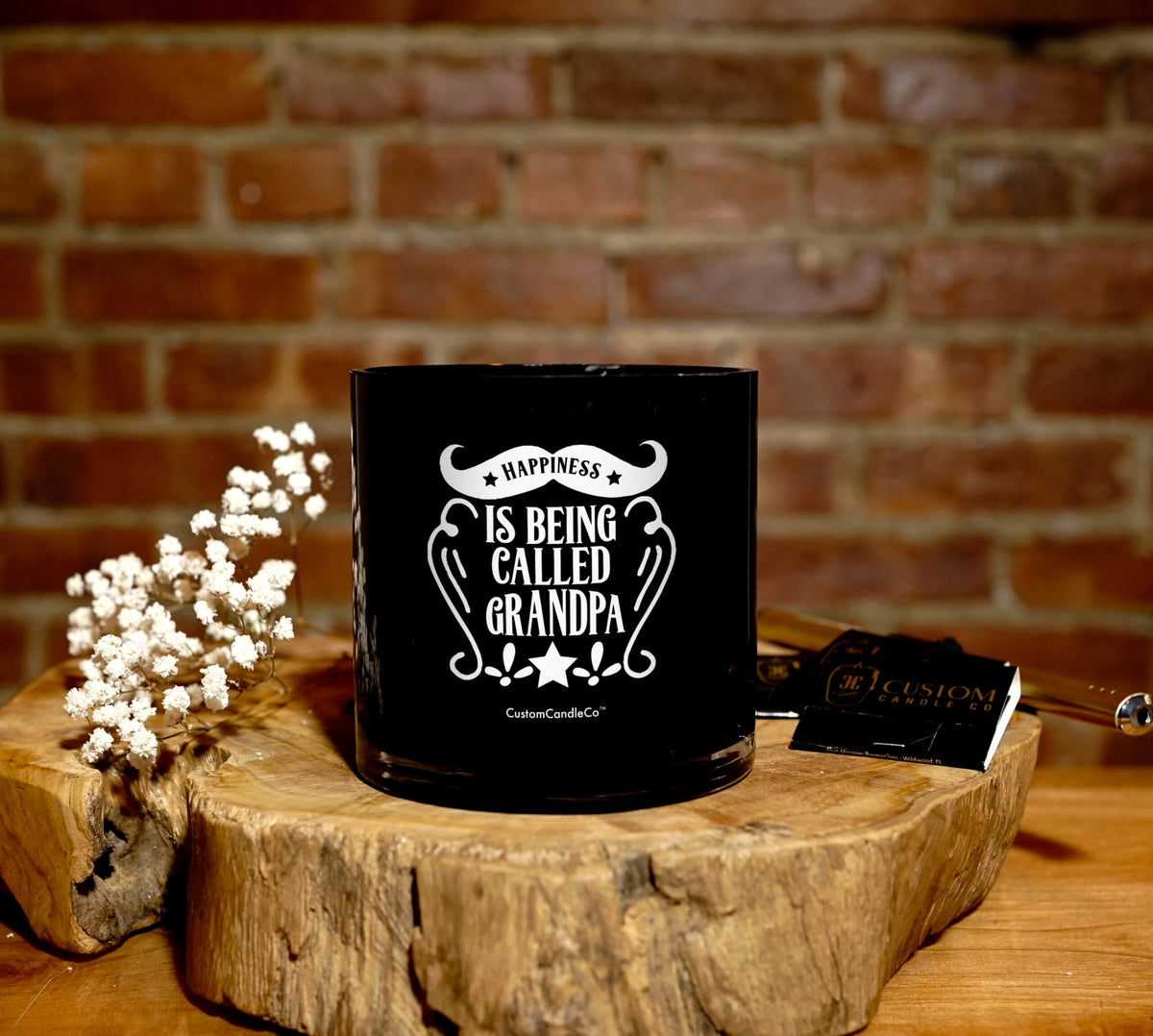 The "Happiness is Being Called Grandpa" Natural Soy Candle sits on a wooden surface, making an ideal gift for grandpa. White flowers and a matchbox lie nearby, all against a brick wall backdrop.