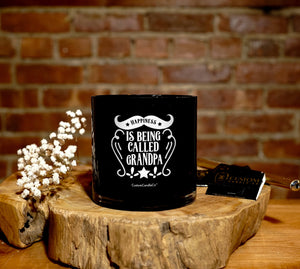 The "Happiness is Being Called Grandpa" Natural Soy Candle sits on a wooden surface, making an ideal gift for grandpa. White flowers and a matchbox lie nearby, all against a brick wall backdrop.