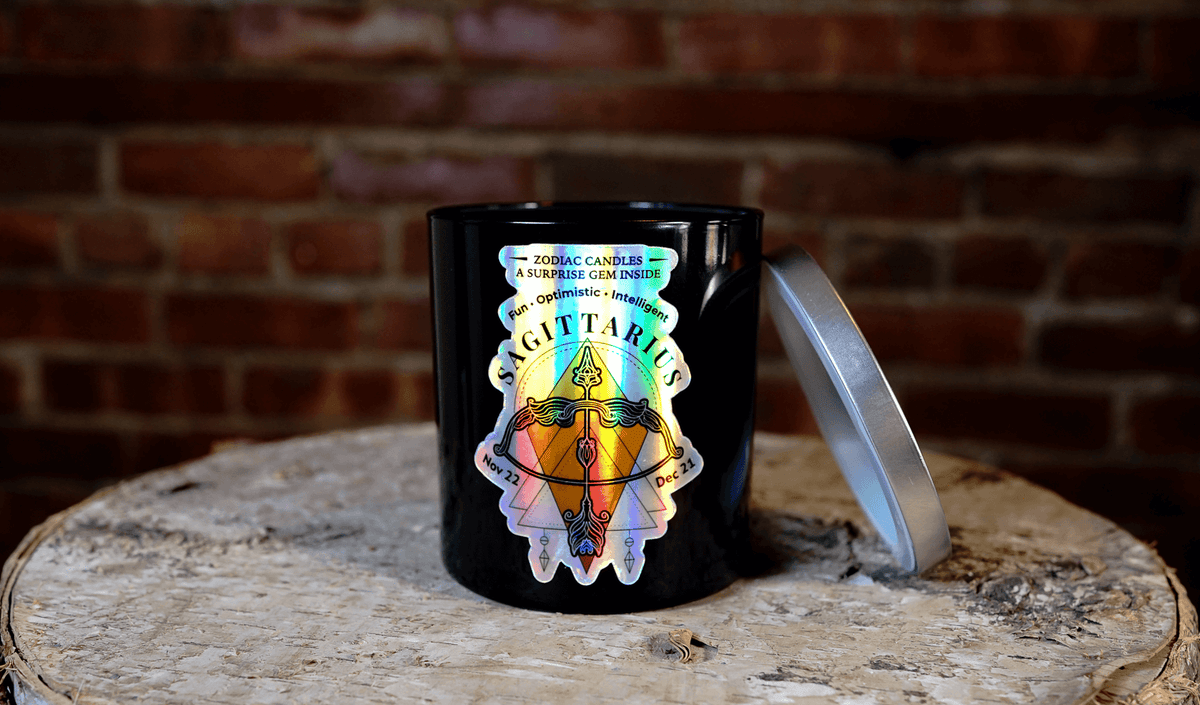 The Zodiac: Sagittarius Candle features a black jar with zodiac-themed artwork and a silver lid, creating a striking celestial accent on any wooden surface against a brick wall.