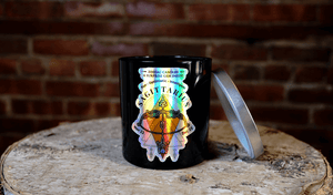 The Zodiac: Sagittarius Candle features a black jar with zodiac-themed artwork and a silver lid, creating a striking celestial accent on any wooden surface against a brick wall.