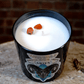 The Zodiac: Aries Candle is a black soy jar labeled "Aries," with two red stones on white wax, displayed on wood against a brick wall.