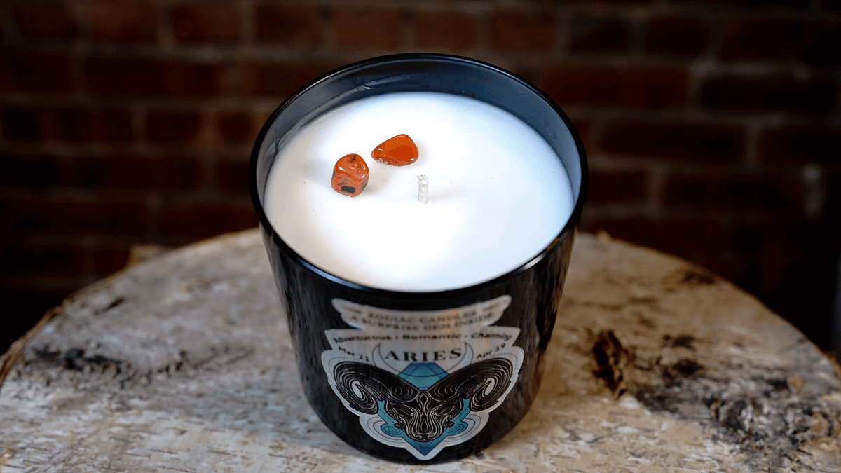 The Zodiac: Aries Candle is a black soy jar labeled "Aries," with two red stones on white wax, displayed on wood against a brick wall.