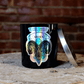 The Zodiac: Aries Candle, featuring a black jar with a metallic label and silver lid, rests on a wooden surface by a brick wall—an ideal soy zodiac candle for astrology enthusiasts.