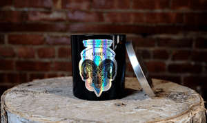 The Zodiac: Aries Candle, featuring a black jar with a metallic label and silver lid, rests on a wooden surface by a brick wall—an ideal soy zodiac candle for astrology enthusiasts.