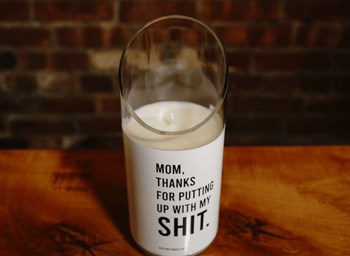A Thanks for Puttin’ Up with My Shit soy candle in a recycled wine bottle holder rests on wood, its cheeky label displayed. A brick wall appears in the background.