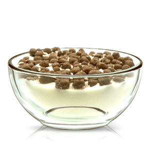 A clear reusable cereal bowl holds a Cereal Puffy Coco Candle, designed to look like white yogurt topped with small round chocolate cereal pieces, set against a white background.