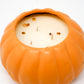 The Ceramic Pumpkin Vessel Candle features a round, orange, pumpkin-shaped ceramic jar with three wicks and natural white soy wax, decorated with a few brown seeds or debris on top.