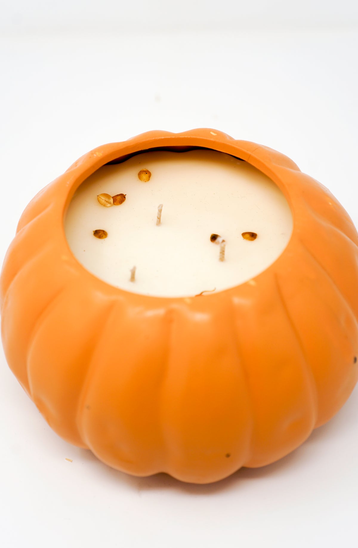 The Ceramic Pumpkin Vessel Candle features a round, orange, pumpkin-shaped ceramic jar with three wicks and natural white soy wax, decorated with a few brown seeds or debris on top.