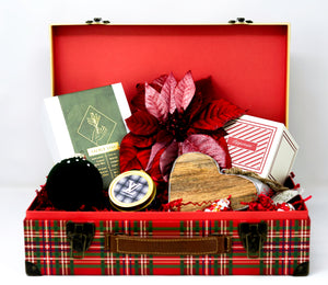Christmas Gift Baskets: Plaid-patterned boxes filled with assorted items such as a book, heart-shaped wooden decor, poinsettia flower, scented candles, and other small gifts on red shredded paper—ideal for holiday gifting.