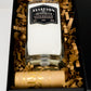 A Recycled American Gin Candle in a black gift box with brown crinkle paper, topped with a wooden cap and complemented by a natural soy candle for an elegant touch.