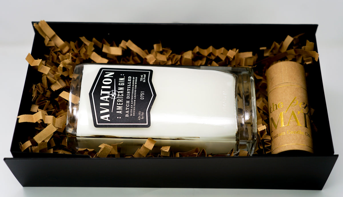 The Recycled American Gin Candle, a natural soy candle in a glass labeled "Aviation American Gin," comes in a black box with brown paper filler beside a small cylindrical container—an eco-friendly accent for any room.