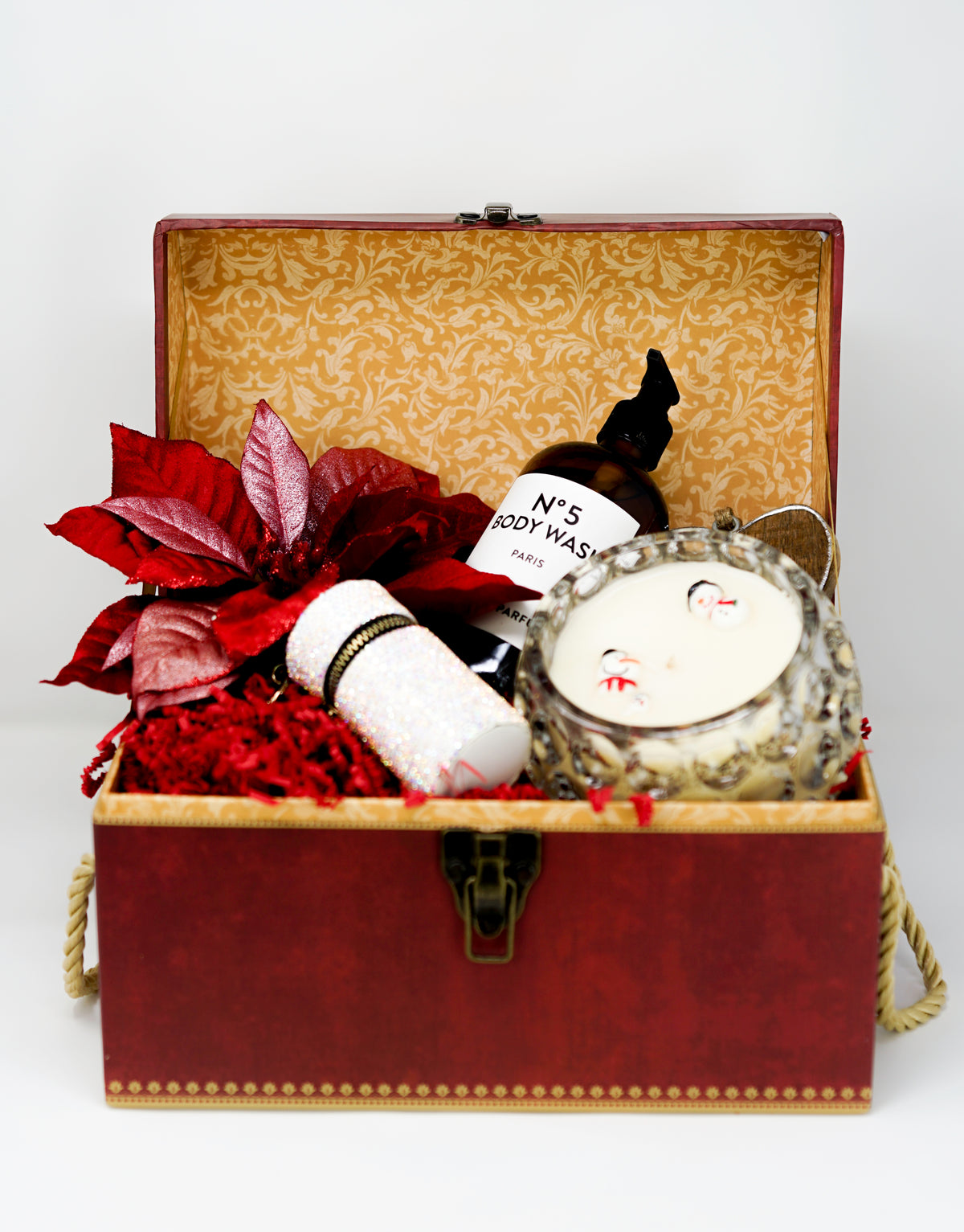An open red gift box from Christmas Gift Baskets with body wash, a scented candle, a glittery cylinder, red leaves, and shredded paper—ideal for holiday gifting.