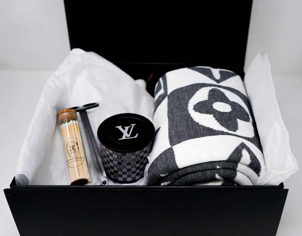 The LV Designer Gift Set includes a rolled cotton throw blanket, a black LV logo candle, a wooden tube, and a silver tool in a black box on white tissue paper—an ideal gift.