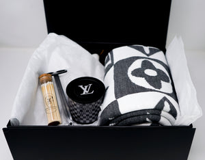 The LV Designer Gift Set includes a rolled cotton throw blanket, a black LV logo candle, a wooden tube, and a silver tool in a black box on white tissue paper—an ideal gift.