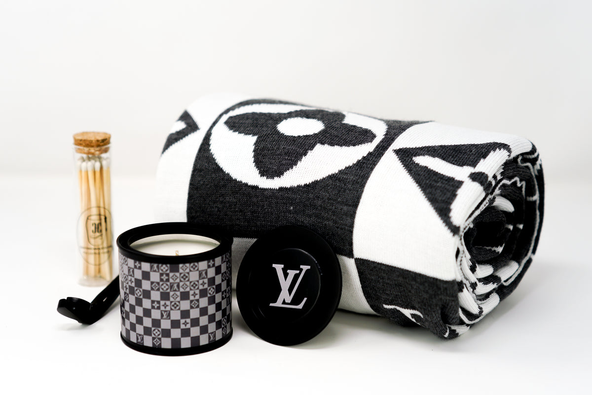 The LV Designer Gift Set features a rolled black and white cotton throw, a sleek black "LV" candle, and a glass jar of wooden matches, all arranged stylishly on a white surface.