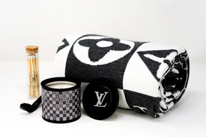 The LV Designer Gift Set features a rolled black and white cotton throw, a sleek black "LV" candle, and a glass jar of wooden matches, all arranged stylishly on a white surface.