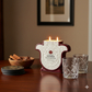Tabletop scene featuring a Recycled Reposado Tequila Candle made from natural soy, two empty etched glasses, a bowl of cinnamon sticks, and stacked coasters.