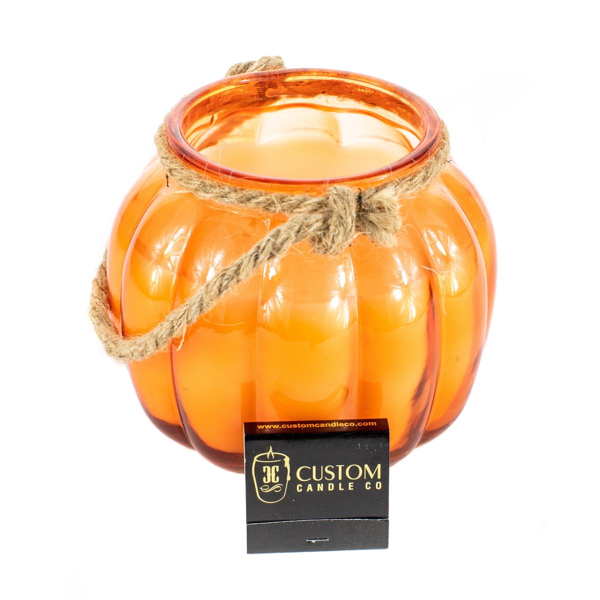 The Glass Pumpkin Lantern features orange glass, a rope handle, and a "Custom Candle Co" label—ideal as a Halloween lantern or as a unique accent to enhance your festive Custom Candle decor.