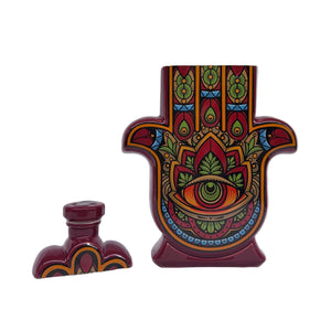 A recycled Reposado Tequila Candle in a ceramic Hamsa hand bottle features colorful patterns and an eye motif, with its matching cap beside it. Perfect for displaying as art or enjoying as a natural soy candle.