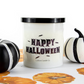 A Happy Halloween Candle in a white frosted jar sits between two black-and-white decorative pumpkins on a surface featuring an orange pumpkin print.