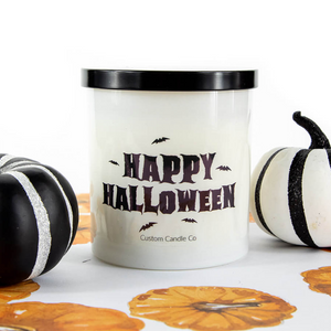 A Happy Halloween Candle in a white frosted jar sits between two black-and-white decorative pumpkins on a surface featuring an orange pumpkin print.