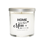 A hand-poured soy candle in a white glass jar with a silver lid. The label says: "Home is where mom is" Custom Candle Co in black decorative text.