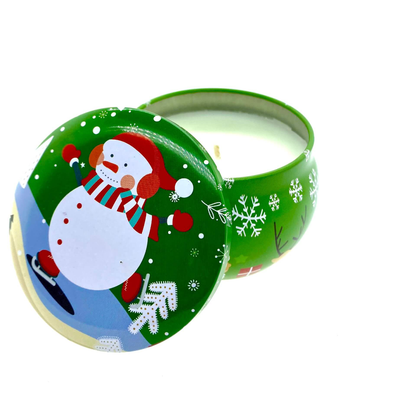 The Green Snowman Holiday Tin Candle – Balsam Scent features cheerful snowman and snowflake designs on the lid. Partially open, it reveals white soy wax, perfect for filling your home with festive fragrance.