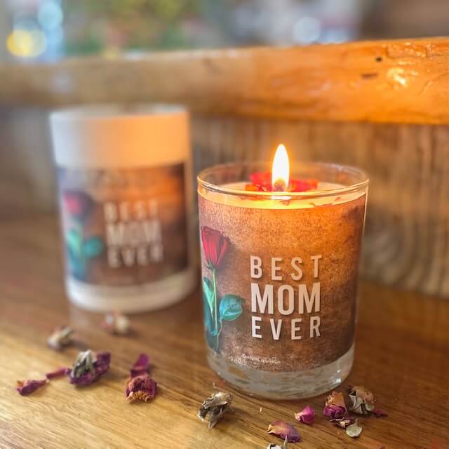 A lit Best Mom Ever quoted candle with box – Amber Scent made of soy wax sits in a glass holder on wood with dried flower petals; another unlit amber-scented candle is visible in the background.