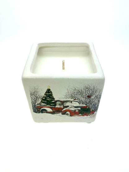The Mini Square Christmas Candle (Snow Design) features a holiday sparkle scent and comes in a ceramic holder with a winter scene—vintage red trucks, snow-covered trees, and bare branches—perfect for your Christmas décor.