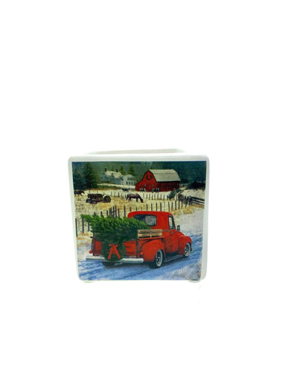 The Mini Square Christmas Candle (Red Truck Design) features a red truck with a Christmas tree on a snowy road, plus a red barn and trees—perfect for your home. Enjoy its natural soy wax and festive Holiday Sparkle scent!.