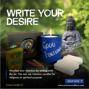 The Intention Blue Candle labeled "Good Fortune," along with chalk, a Buddha statue, and a cloud-shaped decor, sits on greenery. Write your intentions on the candle to enhance spiritual practices or manifest desires.