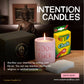 A pink Intention Candle labeled "Good Luck," a Crayola chalk box, Buddha statue, and white candle sit on wood beneath "Intention Candles"—an ideal setup for your spiritual practice.