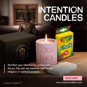 A pink Intention Candle labeled "Good Luck," a Crayola chalk box, Buddha statue, and white candle sit on wood beneath "Intention Candles"—an ideal setup for your spiritual practice.