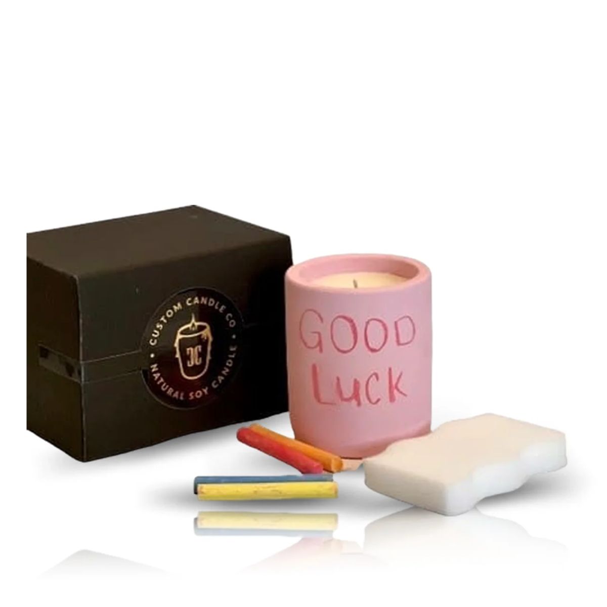 An Intention Pink Candle labeled "GOOD LUCK," three colored chalk sticks, a white eraser, and a black box are arranged on a white background.