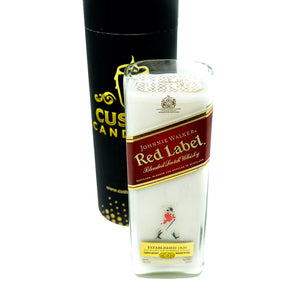 The Recycled Red Label Whiskey Candle is a white recycled candle in a glass holder shaped like a Johnnie Walker Red Label bottle, shown in front of a black cylindrical container.
