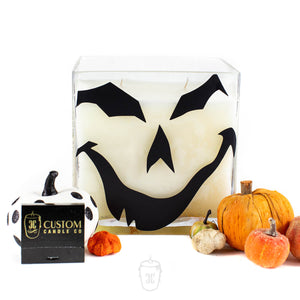 The Jack O’lantern Cube Candle 4 Wick Candle features a square glass design with a black jack-o'-lantern face, surrounded by small pumpkins and a white pumpkin with black dots. Comes in a Custom Candle Co box—perfect for autumn decor and festive vibes.
