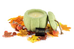 The Green Cement Pumpkin Candle with lid is surrounded by artificial autumn leaves and small decorative apples, placed next to a Custom Candle Co. label.