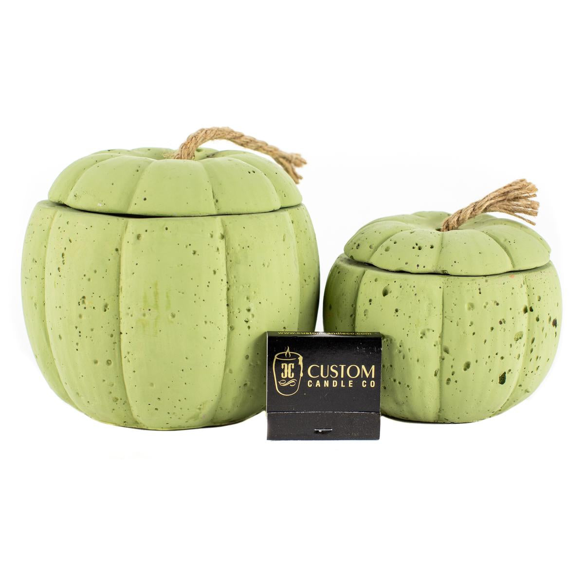 Two Green Cement Pumpkin Candles with rope handles are placed beside a small black "Custom Candle Co." box on a white background.