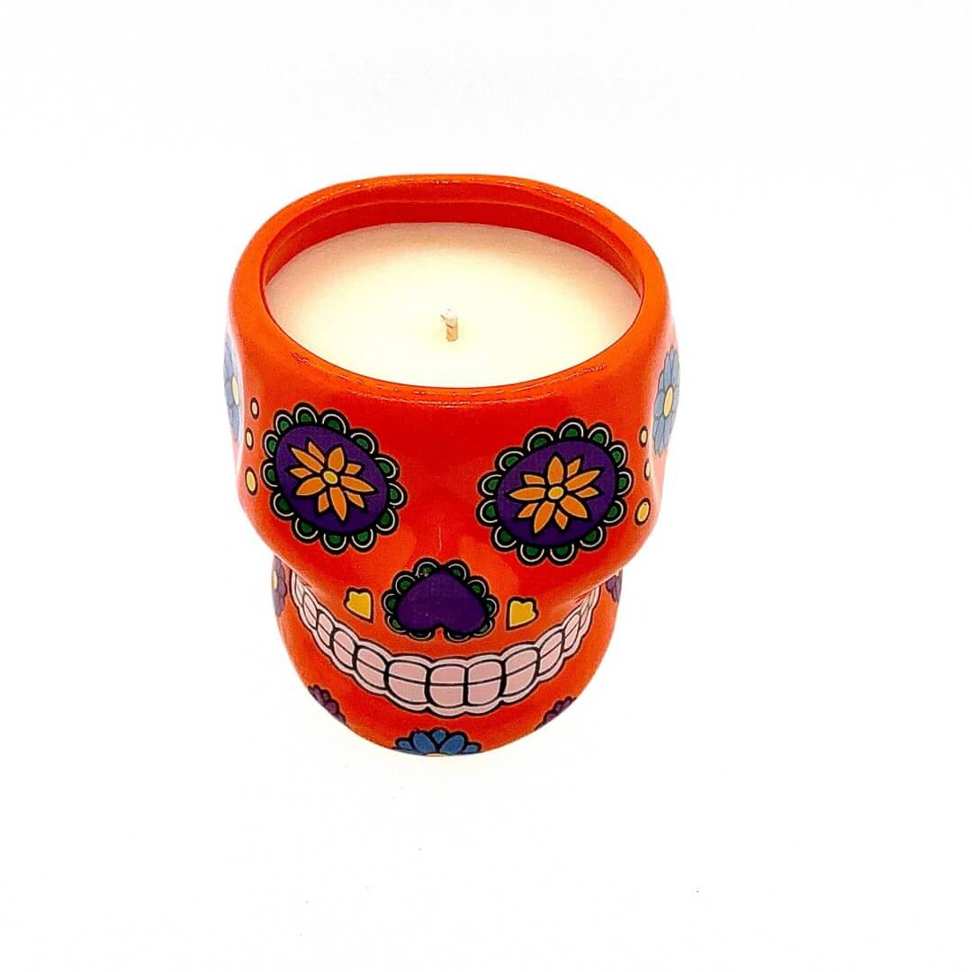 A large orange ceramic skull candle holder with colorful floral designs, featuring a white nutmeg-scented soy wax candle.