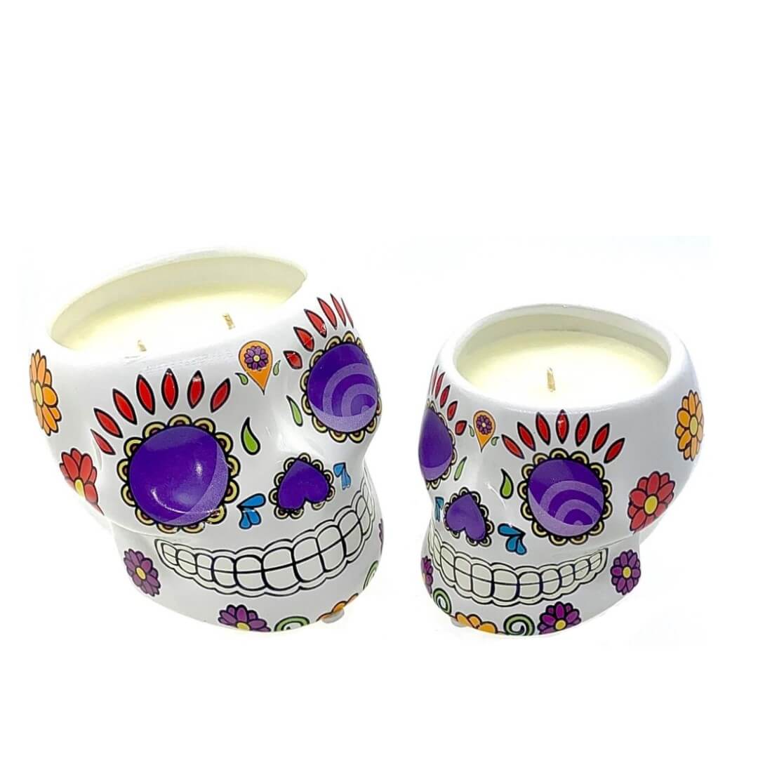 Two Small White Ceramic Skull Candles, nutmeg scented and shaped like decorated sugar skulls with colorful floral patterns and purple accents, are perfect for adding vibrant flair to your space.